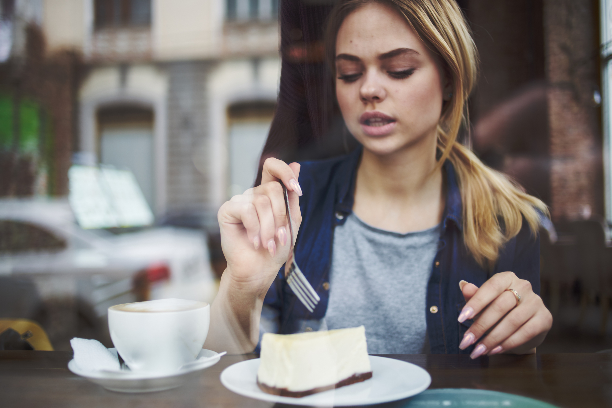 A young woman with blonde hair in a ponytail is sitting at a café table. She is holding a fork and is about to eat a slice of cheesecake on a white plate. There's a cup of coffee with a napkin next to her plate. The background shows a blurred view of buildings.