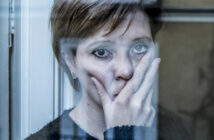 A woman with short hair stands behind a window, her hand covering part of her face. She looks directly at the camera with a somber expression. The window frame partially obscures her, and the scene has a blue-tinted, reflective light.