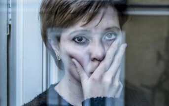 A woman with short hair stands behind a window, her hand covering part of her face. She looks directly at the camera with a somber expression. The window frame partially obscures her, and the scene has a blue-tinted, reflective light.