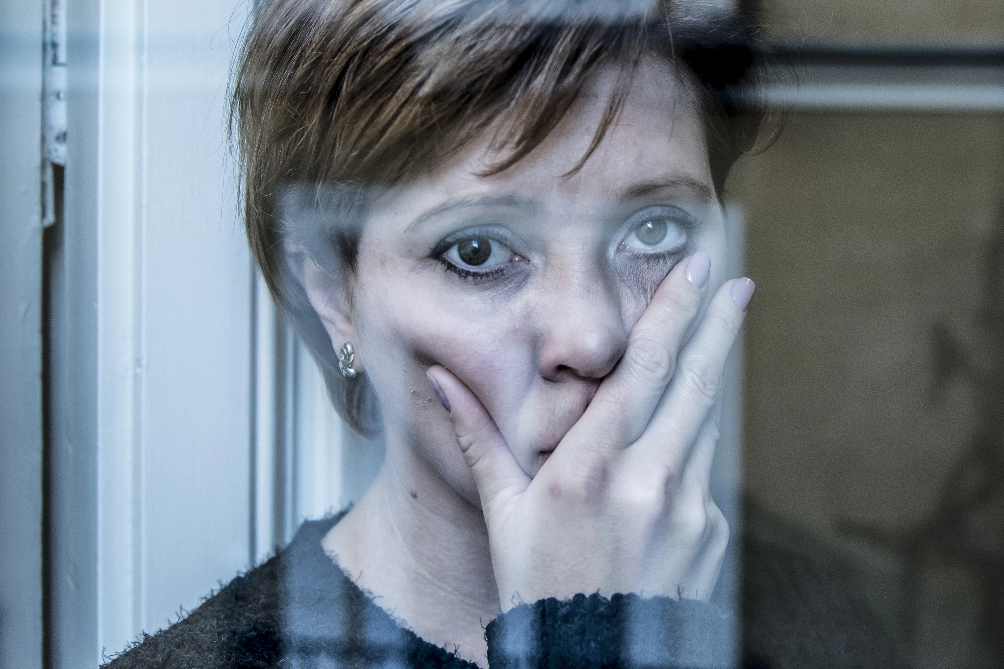 A woman with short hair stands behind a window, her hand covering part of her face. She looks directly at the camera with a somber expression. The window frame partially obscures her, and the scene has a blue-tinted, reflective light.