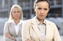 Two businesswomen standing outdoors, both wearing light-colored blazers. The woman in the foreground has dark hair tied back and is looking seriously at the camera with her arms crossed. The woman in the background has blonde hair and is slightly out of focus.