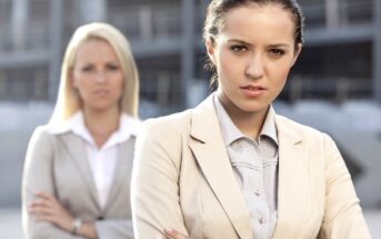 Two businesswomen standing outdoors, both wearing light-colored blazers. The woman in the foreground has dark hair tied back and is looking seriously at the camera with her arms crossed. The woman in the background has blonde hair and is slightly out of focus.