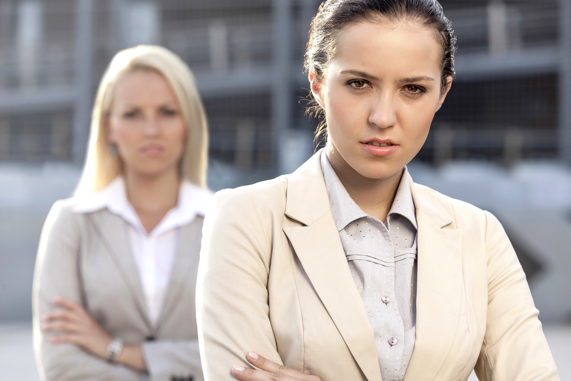 Two businesswomen standing outdoors, both wearing light-colored blazers. The woman in the foreground has dark hair tied back and is looking seriously at the camera with her arms crossed. The woman in the background has blonde hair and is slightly out of focus.