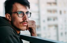 A man with glasses and a mustache wearing a dark hoodie leans on a railing while gazing thoughtfully into the distance. There are blurred building structures in the background, suggesting an urban setting.