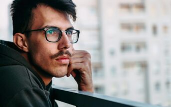 A man with glasses and a mustache wearing a dark hoodie leans on a railing while gazing thoughtfully into the distance. There are blurred building structures in the background, suggesting an urban setting.