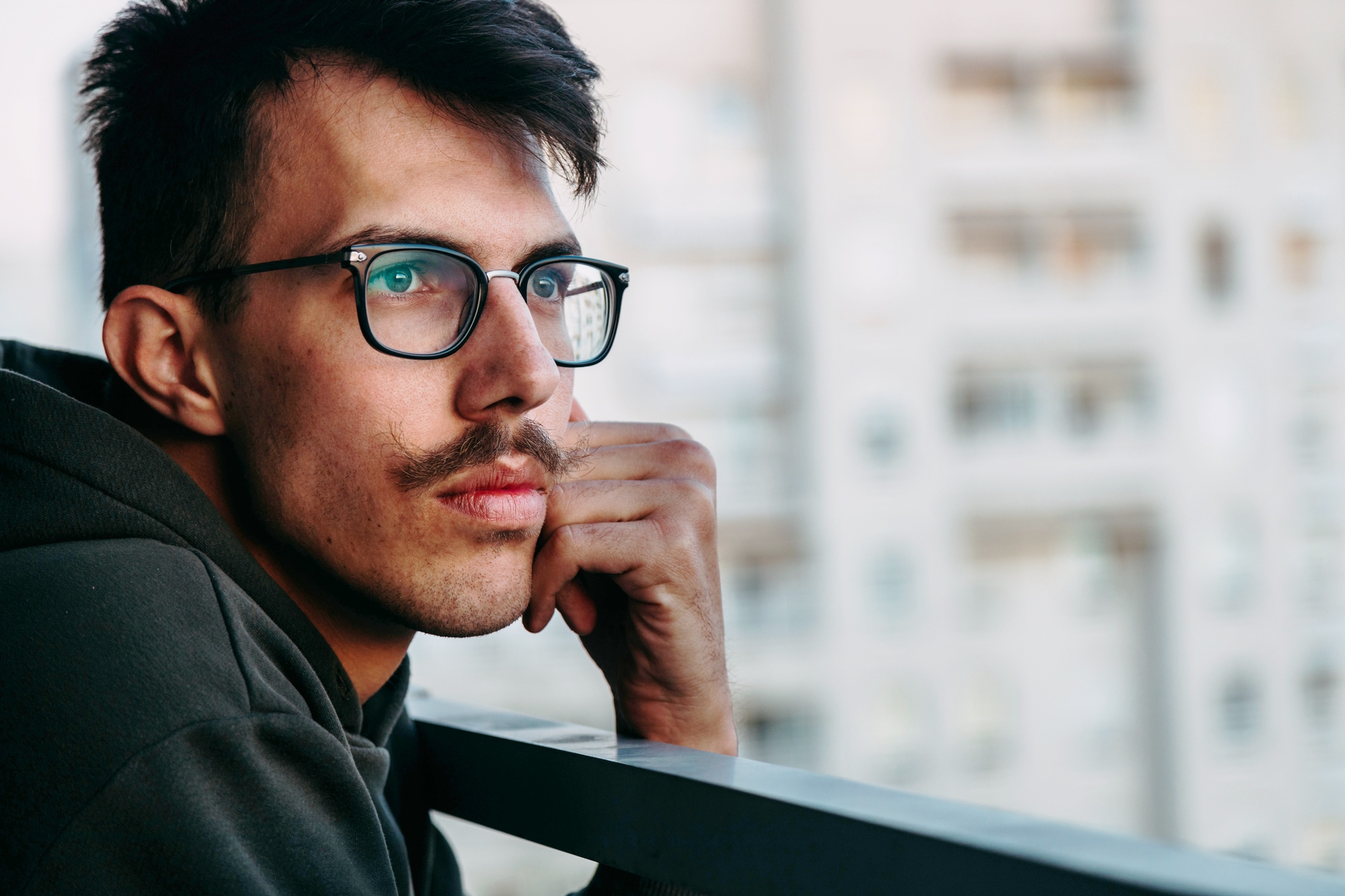 A man with glasses and a mustache wearing a dark hoodie leans on a railing while gazing thoughtfully into the distance. There are blurred building structures in the background, suggesting an urban setting.