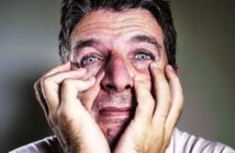 A close-up of a distressed man with short hair and blue eyes, with visible tears and furrowed brows. He is holding his face with both hands, fingers pressing against his cheeks, conveying an intense emotional state. The background is blurred and neutral in color.