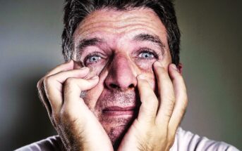 A close-up of a distressed man with short hair and blue eyes, with visible tears and furrowed brows. He is holding his face with both hands, fingers pressing against his cheeks, conveying an intense emotional state. The background is blurred and neutral in color.