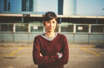 A woman with dark hair tied back stands with her arms crossed, wearing a maroon sweater over a gray shirt. She stands outdoors in an urban setting with a wire fence and modern buildings in the background on a sunny day.