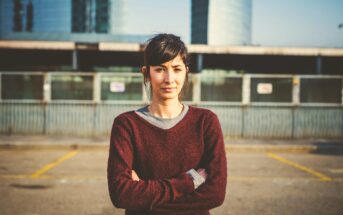 A woman with dark hair tied back stands with her arms crossed, wearing a maroon sweater over a gray shirt. She stands outdoors in an urban setting with a wire fence and modern buildings in the background on a sunny day.