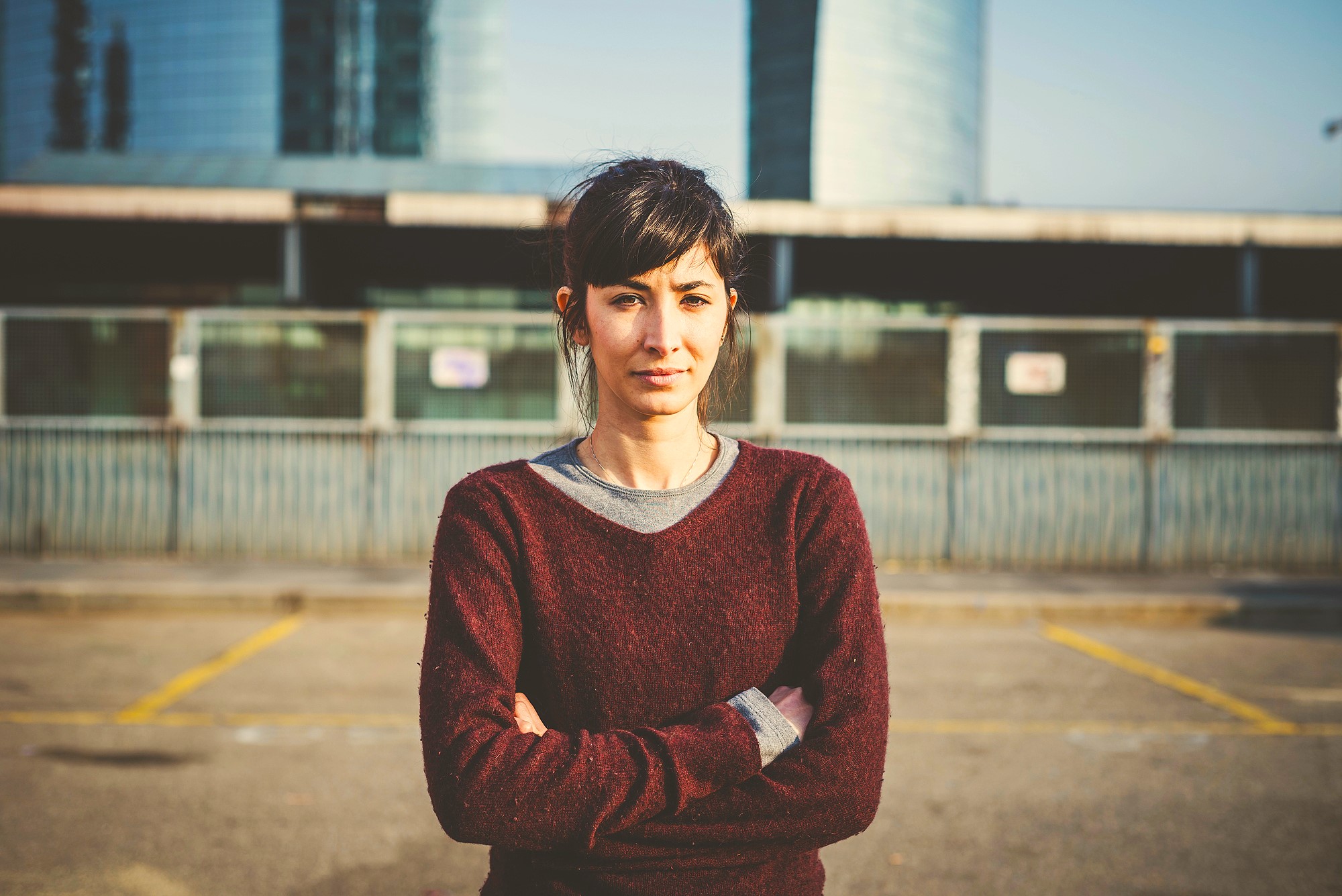 A woman with dark hair tied back stands with her arms crossed, wearing a maroon sweater over a gray shirt. She stands outdoors in an urban setting with a wire fence and modern buildings in the background on a sunny day.