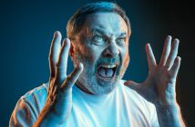 A man with short gray hair and a beard is angrily shouting with his mouth wide open. His hands are raised beside his face, fingers splayed. He is wearing a white shirt and is dramatically lit with blue and orange lighting, set against a dark background.