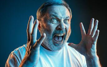 A man with short gray hair and a beard is angrily shouting with his mouth wide open. His hands are raised beside his face, fingers splayed. He is wearing a white shirt and is dramatically lit with blue and orange lighting, set against a dark background.