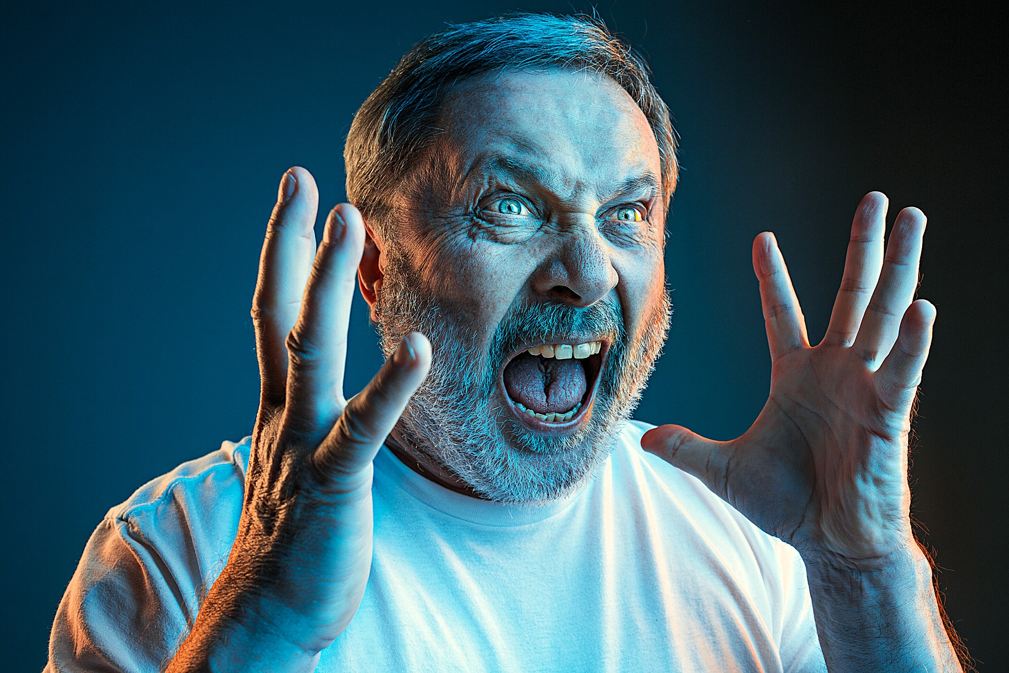 A man with short gray hair and a beard is angrily shouting with his mouth wide open. His hands are raised beside his face, fingers splayed. He is wearing a white shirt and is dramatically lit with blue and orange lighting, set against a dark background.