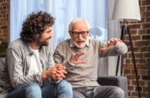 An older man with gray hair and glasses, wearing a gray sweater, animatedly explains something with outstretched hands to a younger man with curly hair and a beard, dressed in a casual gray shirt. They are seated on a couch in a cozy room with a brick wall.