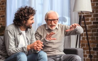 An older man with gray hair and glasses, wearing a gray sweater, animatedly explains something with outstretched hands to a younger man with curly hair and a beard, dressed in a casual gray shirt. They are seated on a couch in a cozy room with a brick wall.