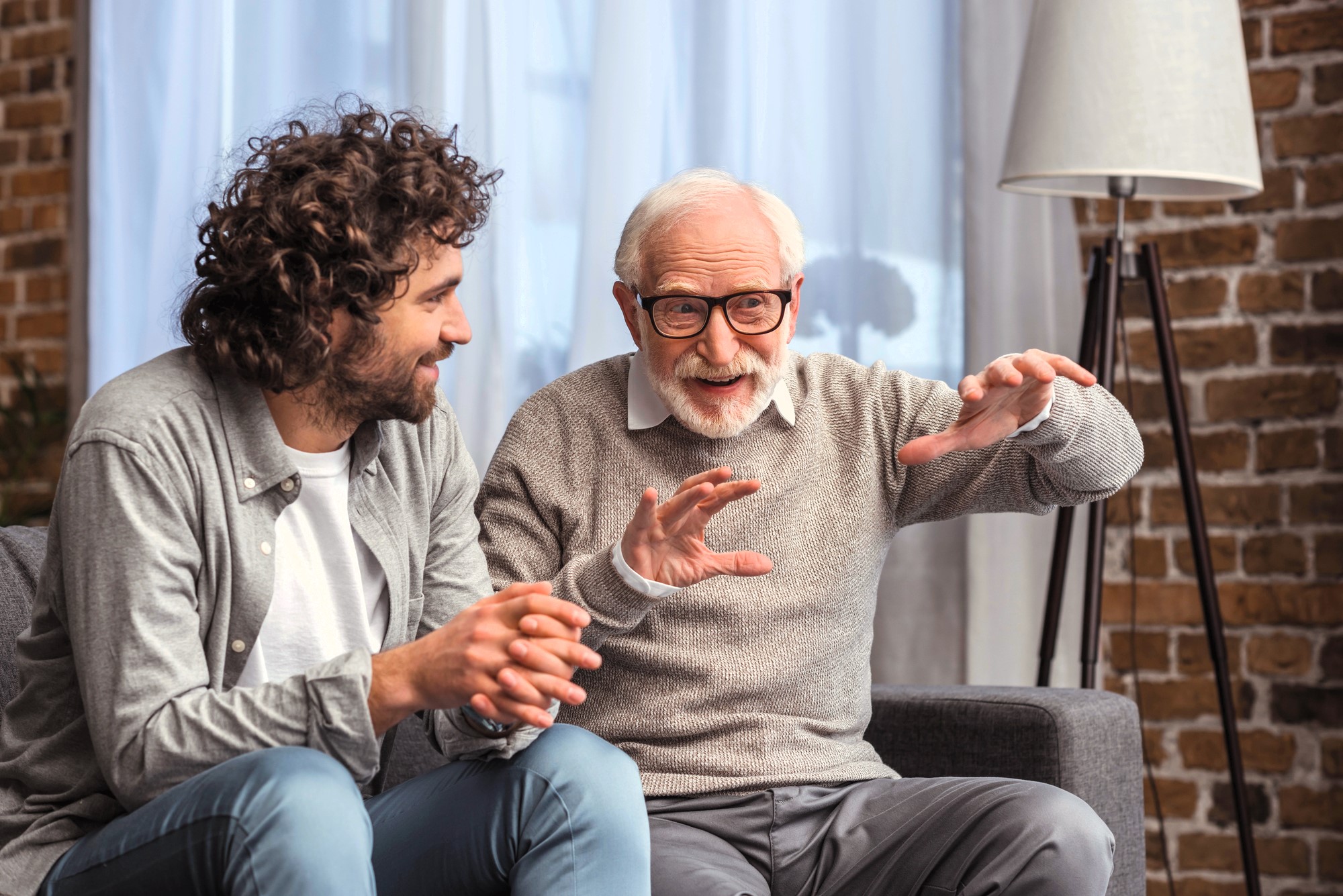 An older man with gray hair and glasses, wearing a gray sweater, animatedly explains something with outstretched hands to a younger man with curly hair and a beard, dressed in a casual gray shirt. They are seated on a couch in a cozy room with a brick wall.