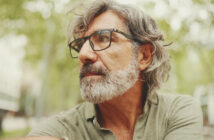 An older man with gray hair and a beard is wearing glasses and a green shirt. He is outdoors with trees and a blurry urban background, looking off to the side thoughtfully.