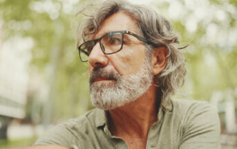 An older man with gray hair and a beard is wearing glasses and a green shirt. He is outdoors with trees and a blurry urban background, looking off to the side thoughtfully.