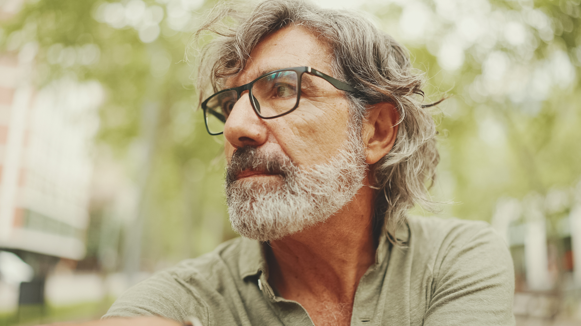 An older man with gray hair and a beard is wearing glasses and a green shirt. He is outdoors with trees and a blurry urban background, looking off to the side thoughtfully.