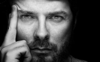 A black and white close-up of a man with a beard gazing intently at the camera. He has one finger touching his temple and appears deep in thought. His serious expression is highlighted by the high contrast of the lighting.