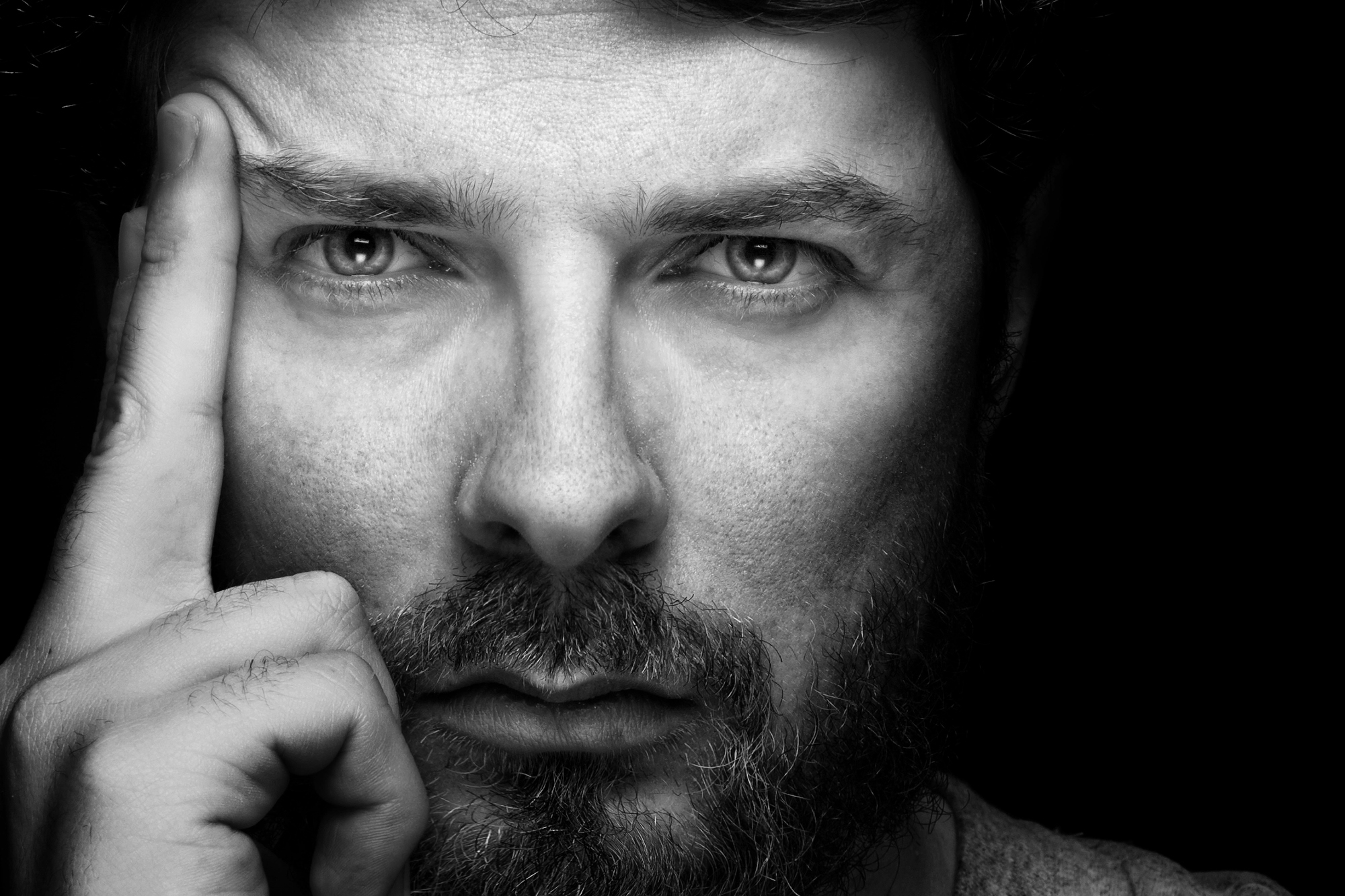 A black and white close-up of a man with a beard gazing intently at the camera. He has one finger touching his temple and appears deep in thought. His serious expression is highlighted by the high contrast of the lighting.