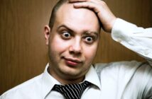 A man wearing a white dress shirt and a loosely knotted black and white striped tie is standing in front of a plain wooden background. He has a surprised expression on his face, with wide eyes and raised eyebrows, while holding his head with one hand.