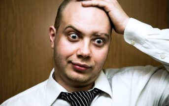 A man wearing a white dress shirt and a loosely knotted black and white striped tie is standing in front of a plain wooden background. He has a surprised expression on his face, with wide eyes and raised eyebrows, while holding his head with one hand.