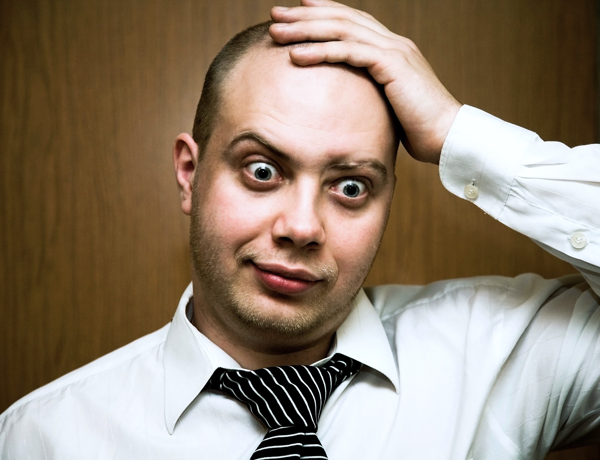 A man wearing a white dress shirt and a loosely knotted black and white striped tie is standing in front of a plain wooden background. He has a surprised expression on his face, with wide eyes and raised eyebrows, while holding his head with one hand.