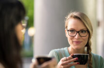 Two women enjoying coffee together outdoors. One woman, with glasses and a braid, holds a black cup and smiles at the other woman, who is blurred in the foreground. The background features a green, blurred natural setting.