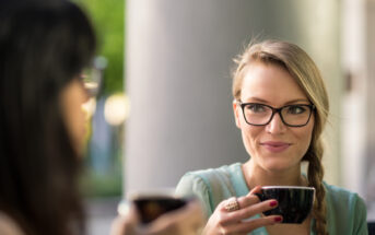 Two women enjoying coffee together outdoors. One woman, with glasses and a braid, holds a black cup and smiles at the other woman, who is blurred in the foreground. The background features a green, blurred natural setting.