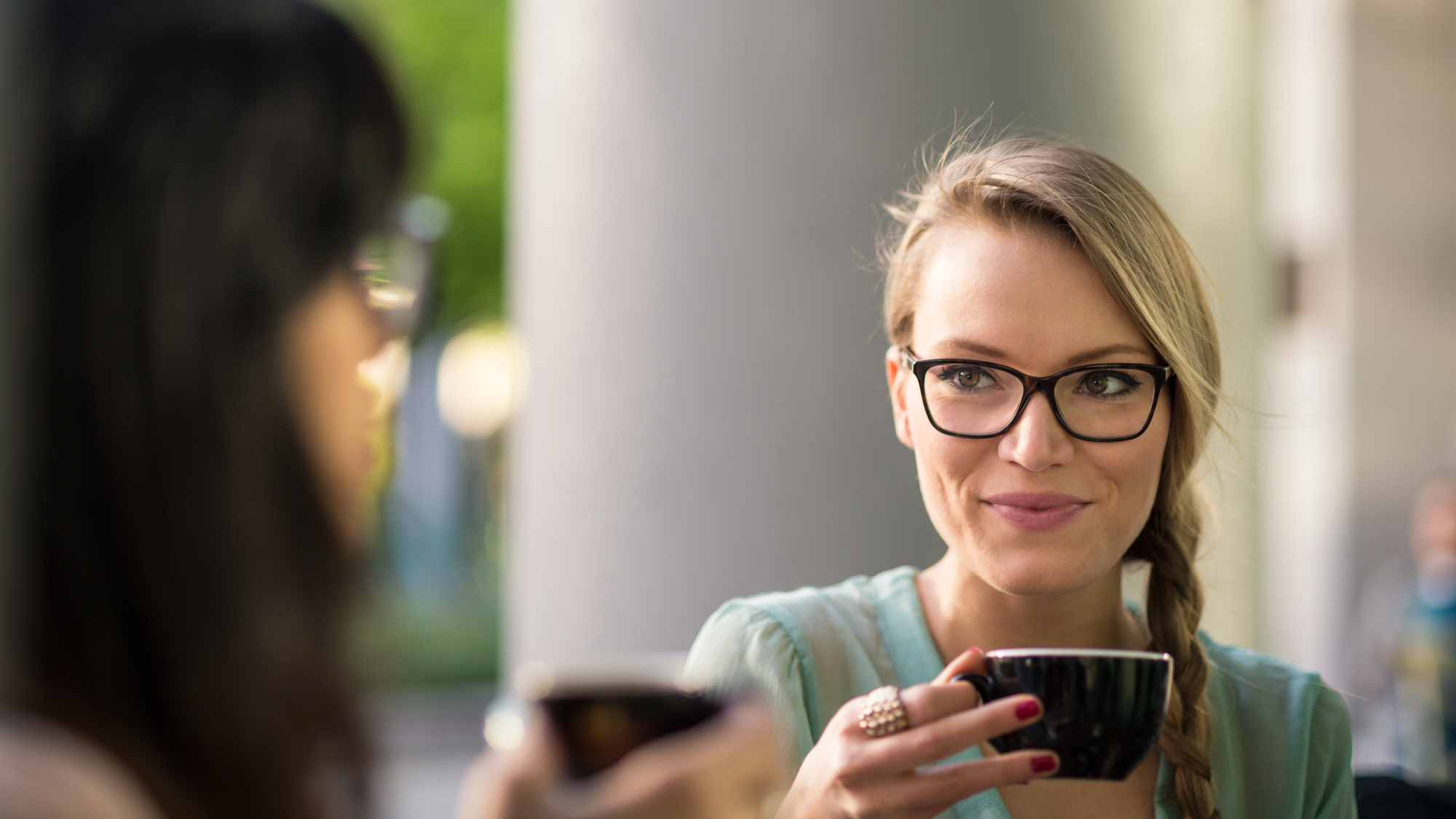 Two women enjoying coffee together outdoors. One woman, with glasses and a braid, holds a black cup and smiles at the other woman, who is blurred in the foreground. The background features a green, blurred natural setting.