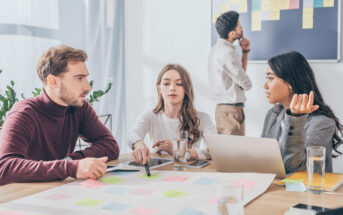 Three individuals are sitting at a table with colorful sticky notes and a laptop, engaged in a discussion. A fourth person in the background is placing notes on a board. Glasses of water are on the table, and there are plants in the room.