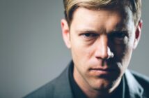 Close-up portrait of a stern-looking man with blond hair, wearing a dark jacket. The background is a smooth gradient, transitioning from light on the left to darker on the right. The man is looking directly into the camera with a serious expression.