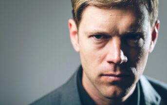 Close-up portrait of a stern-looking man with blond hair, wearing a dark jacket. The background is a smooth gradient, transitioning from light on the left to darker on the right. The man is looking directly into the camera with a serious expression.