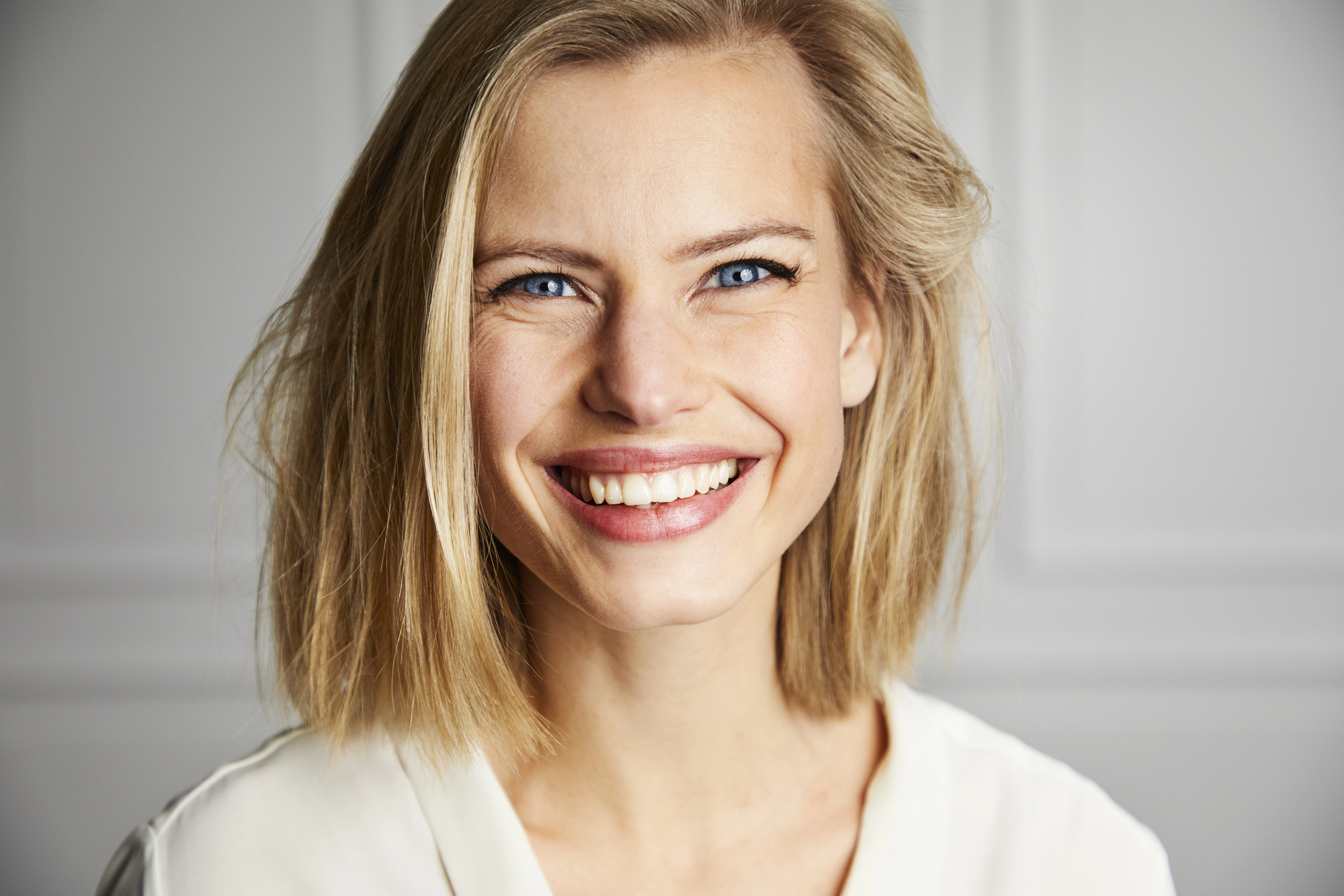 A person with shoulder-length blonde hair smiles brightly at the camera. They are wearing a white shirt and have a light complexion, blue eyes, and slightly tousled hair. The background is softly blurred, likely an indoor setting with white walls.