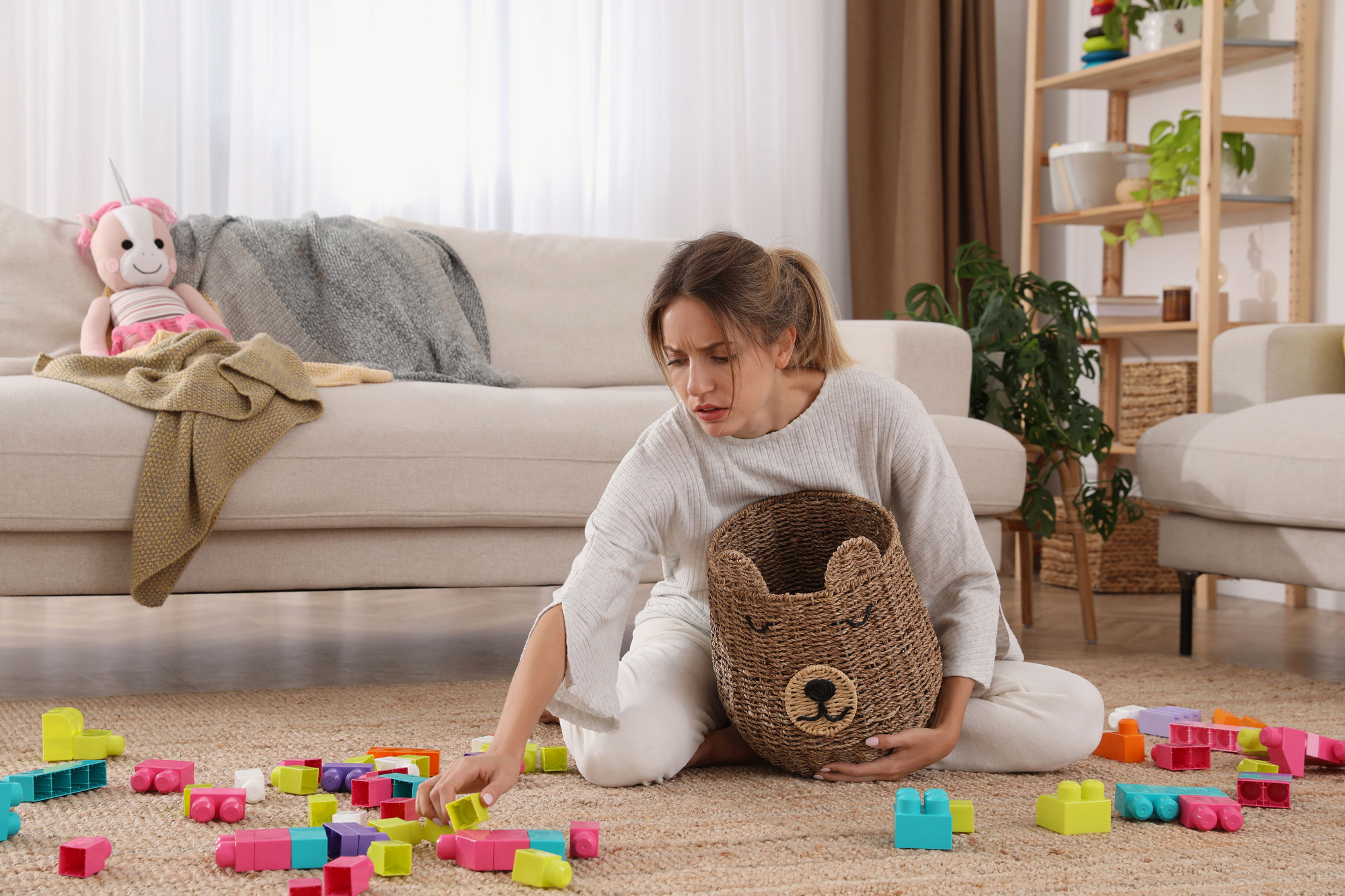 A woman sits on the floor, holding a basket shaped like a bear's face, picking up colorful toy building blocks scattered around her. Behind her is a beige sofa with a gray blanket and a pink stuffed animal, set in a living room with a plant and shelves.