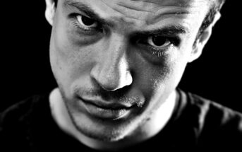 Close-up black and white image of a man with a serious, intense expression. The lighting highlights his facial features, creating strong contrasts and shadows, particularly around his eyes and forehead. He wears a dark shirt, and the background is pitch black.