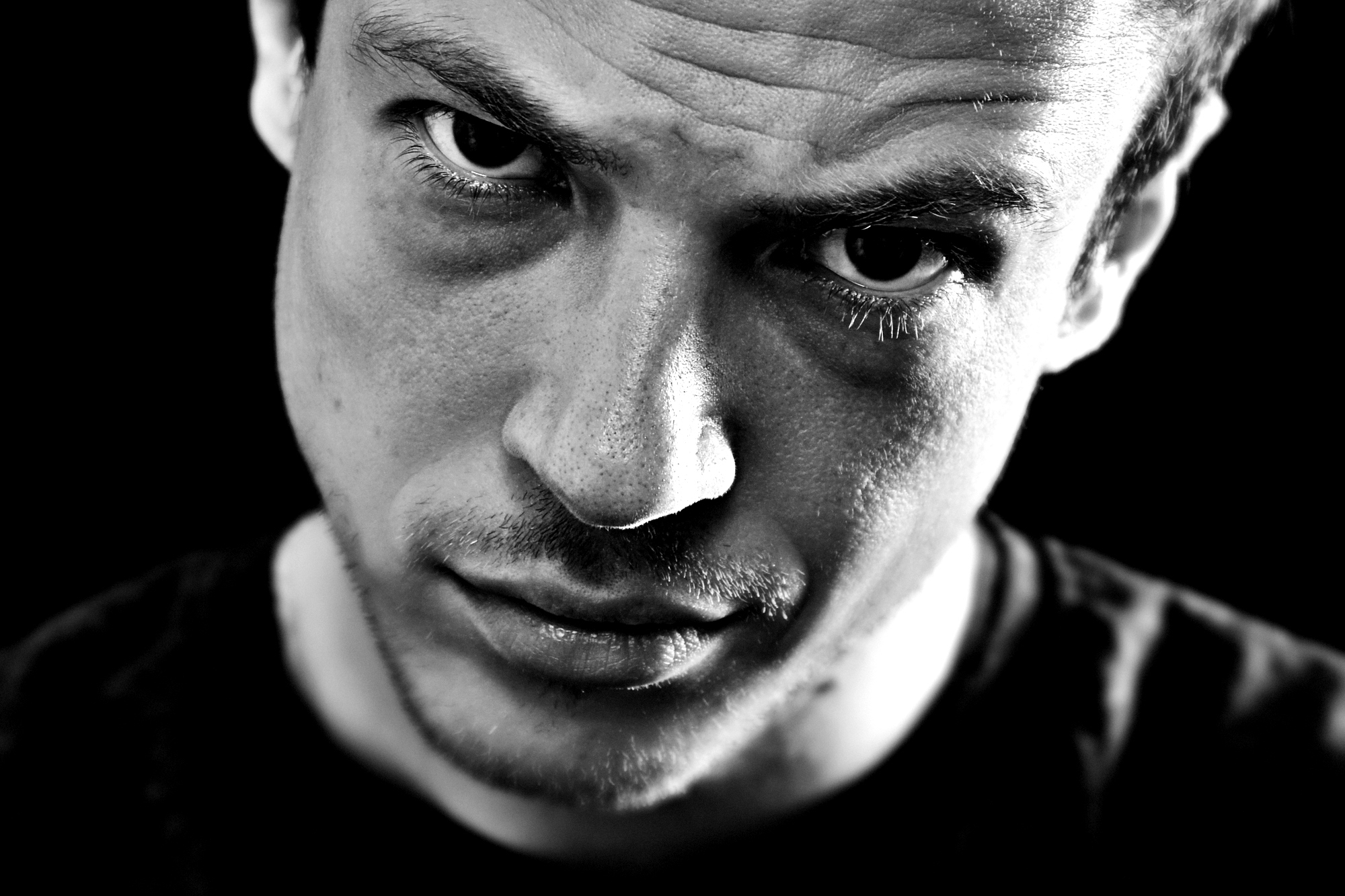 Close-up black and white image of a man with a serious, intense expression. The lighting highlights his facial features, creating strong contrasts and shadows, particularly around his eyes and forehead. He wears a dark shirt, and the background is pitch black.