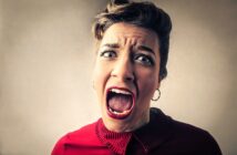 A woman with a pin-up hairstyle and bold makeup is shouting or screaming with her mouth wide open and eyes widened. She is wearing a red blouse with a ruffled collar and has a stressed expression. The background is a plain, muted color.