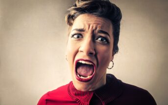 A woman with a pin-up hairstyle and bold makeup is shouting or screaming with her mouth wide open and eyes widened. She is wearing a red blouse with a ruffled collar and has a stressed expression. The background is a plain, muted color.