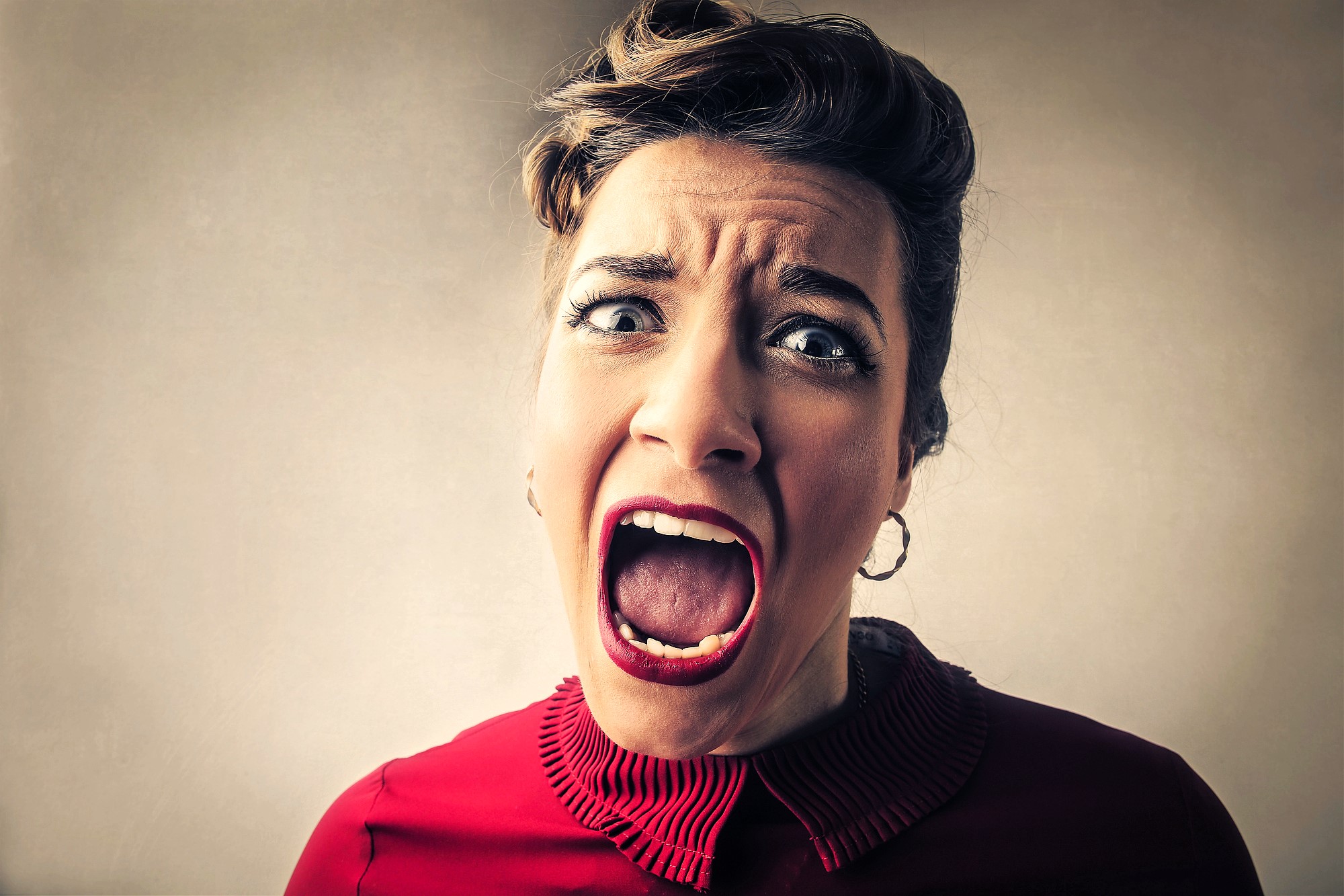 A woman with a pin-up hairstyle and bold makeup is shouting or screaming with her mouth wide open and eyes widened. She is wearing a red blouse with a ruffled collar and has a stressed expression. The background is a plain, muted color.