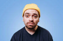 A man with a serious expression wears a yellow beanie and a dark blue T-shirt. He has a short beard and mustache, and his eyes are slightly squinted. The background is a solid light blue color.