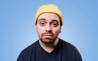 A man with a serious expression wears a yellow beanie and a dark blue T-shirt. He has a short beard and mustache, and his eyes are slightly squinted. The background is a solid light blue color.