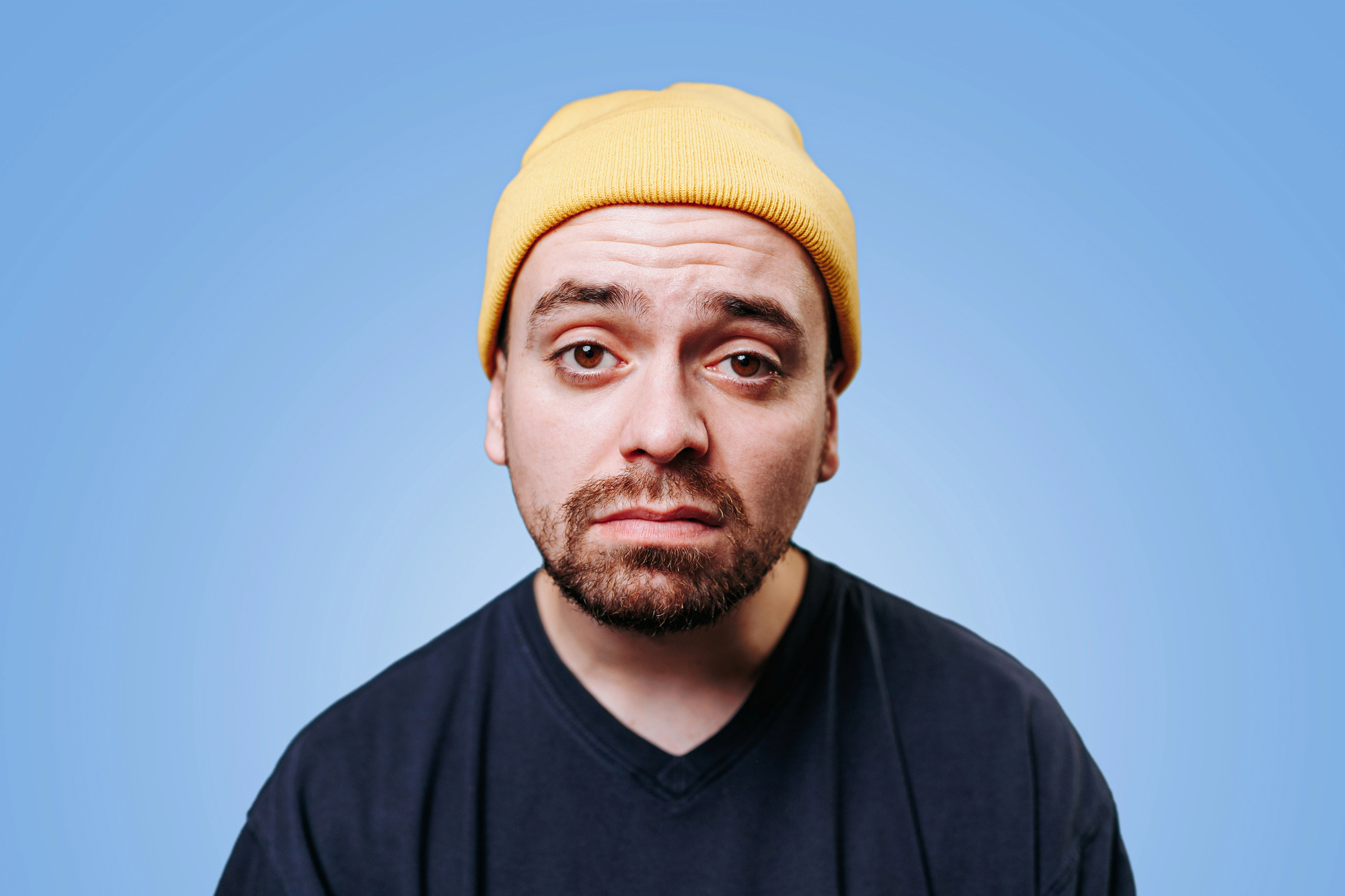 A man with a serious expression wears a yellow beanie and a dark blue T-shirt. He has a short beard and mustache, and his eyes are slightly squinted. The background is a solid light blue color.