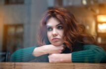 A woman with long, wavy auburn hair wears a green sweater and rests her chin on her crossed arms on a windowsill. She looks thoughtfully out the window with a neutral expression. The background is softly blurred with warm lighting.