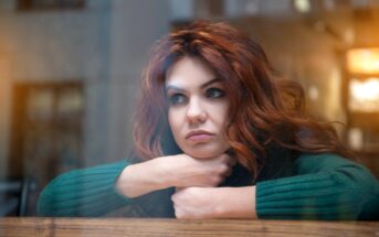 A woman with long, wavy auburn hair wears a green sweater and rests her chin on her crossed arms on a windowsill. She looks thoughtfully out the window with a neutral expression. The background is softly blurred with warm lighting.