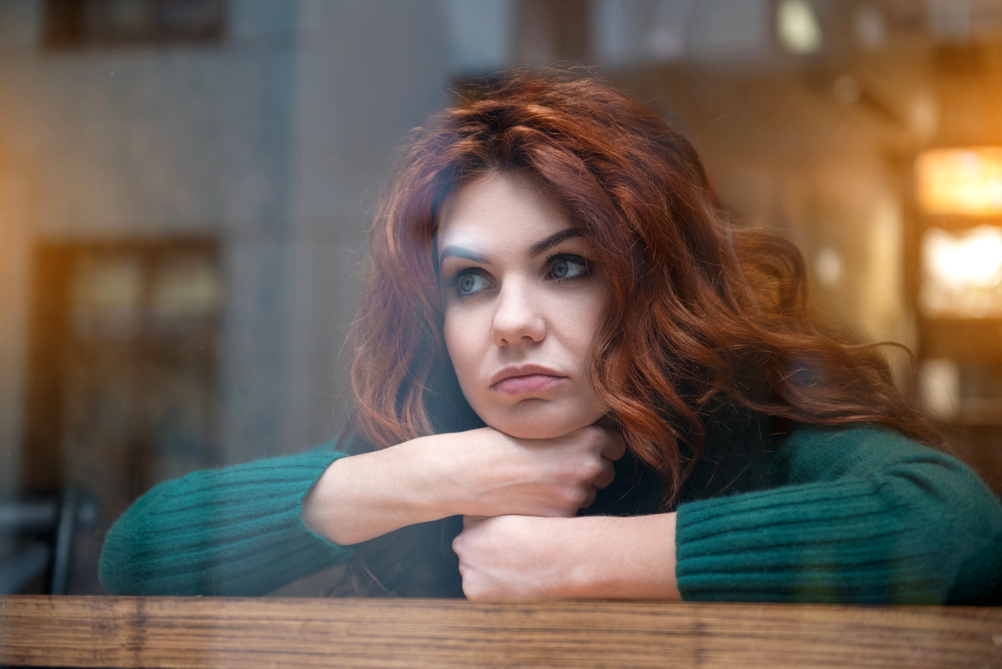 A woman with long, wavy auburn hair wears a green sweater and rests her chin on her crossed arms on a windowsill. She looks thoughtfully out the window with a neutral expression. The background is softly blurred with warm lighting.