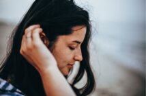 Close-up of a young woman with long dark hair, tilting her head slightly down while gently touching her ear with one hand. She has a serene expression and appears to be outdoors, with a blurred background suggesting a beach or seaside location.