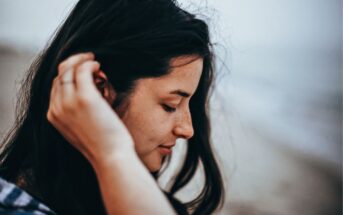 Close-up of a young woman with long dark hair, tilting her head slightly down while gently touching her ear with one hand. She has a serene expression and appears to be outdoors, with a blurred background suggesting a beach or seaside location.